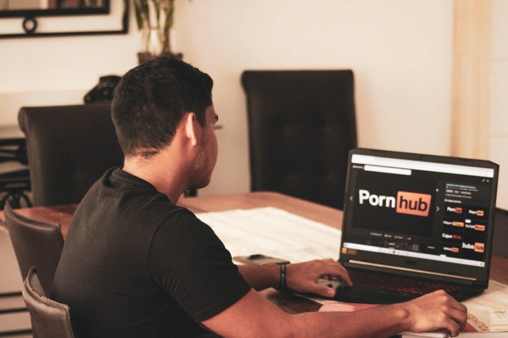 A man wearing a black t-shirt viewing the pornhub website on his laptop in his office 